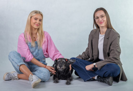 Na zdjęciu widoczne są dwie osoby siedzące na podłodze na jednolitym, jasnym tle. Między nimi znajduje się mały czarny pies. Obie osoby mają swobodną, nieformalną postawę i delikatnie głaszczą zwierzę. Scena ma spokojny, przyjazny charakter